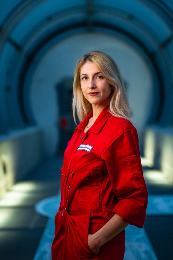 Osoba w czerwonym kombinezonie w industrialnym otoczeniu z metalowym tunelem w tle.