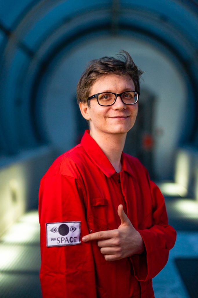 Osoba w czerwonym kombinezonie w tunelu wskazuje na naszywkę z napisem "INN SPACE".