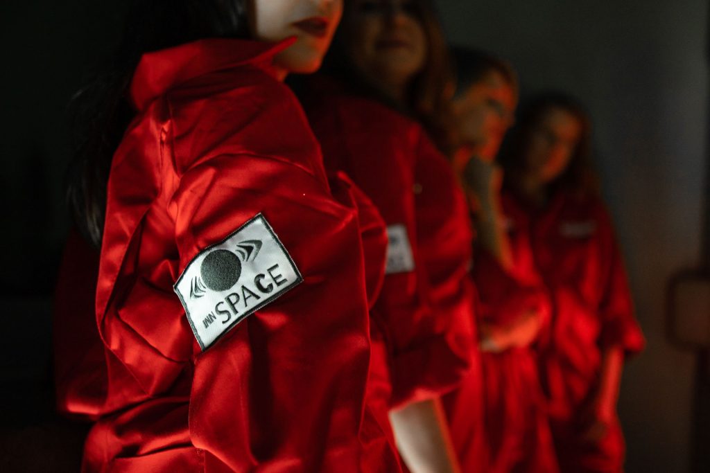 Osoby w czerwonych kombinezonach z logo „INN SPACE”, częściowo w cieniu.
