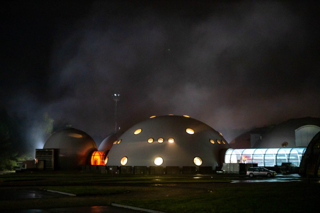 Futurystyczne, kopulaste budynki nocą, z okrągłymi oknami i mgłą w tle.