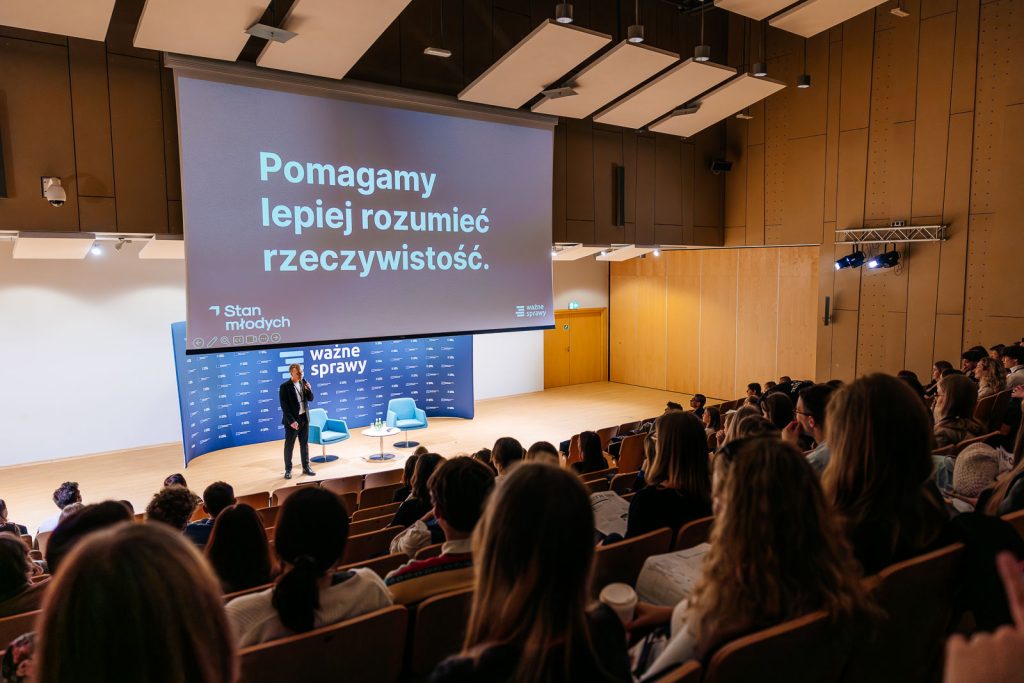Sala wykładowa z dużym ekranem z tekstem, prelegent na scenie z publicznością.