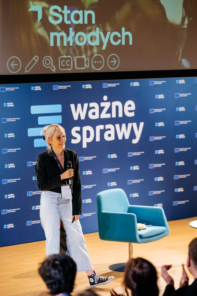 Osoba z mikrofonem na scenie, niebieskie tło z napisem "ważne sprawy", obok krzesło.