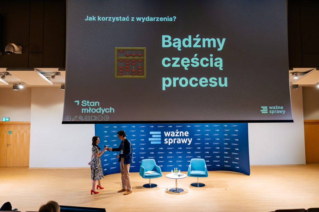 Dwie osoby na scenie z dużym ekranem, niebieskimi fotelami i stolikiem z wodą w tle.