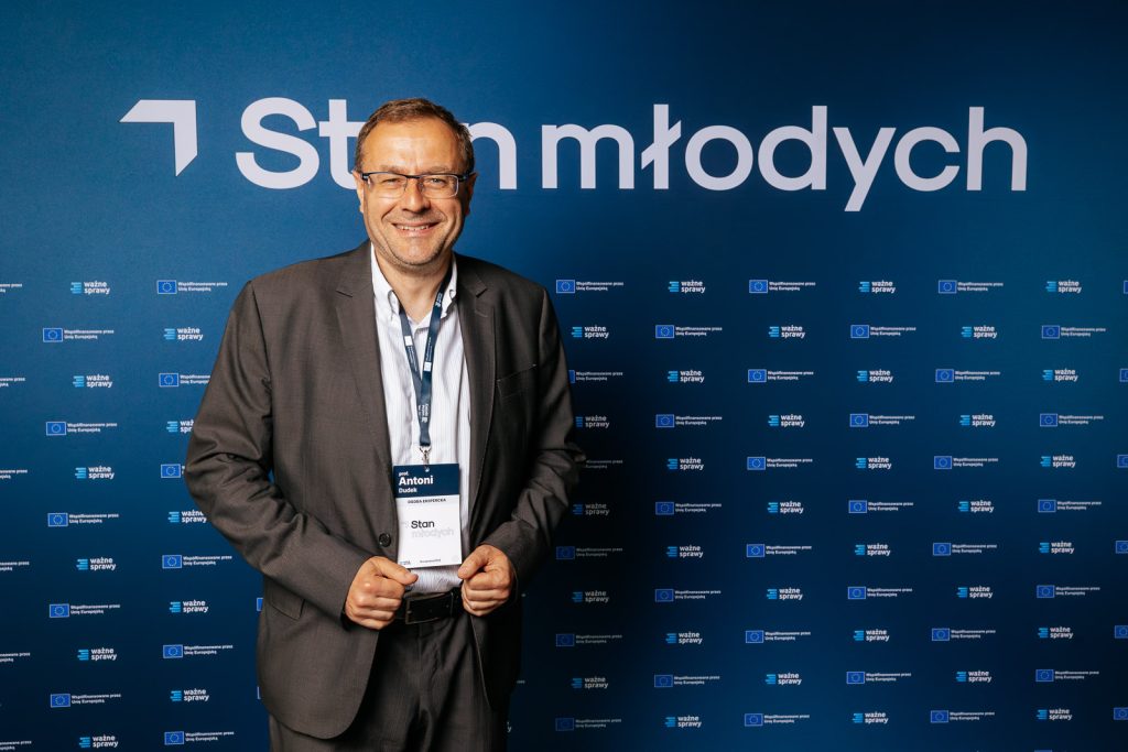 Mężczyzna w garniturze przed ścianką "Stan młodych" z logotypami UE, trzyma identyfikator.