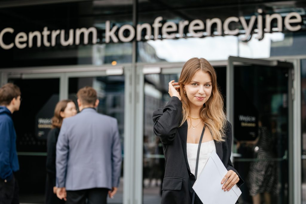 Kobieta z dokumentami przed wejściem do centrum konferencyjnego, ludzie w tle.