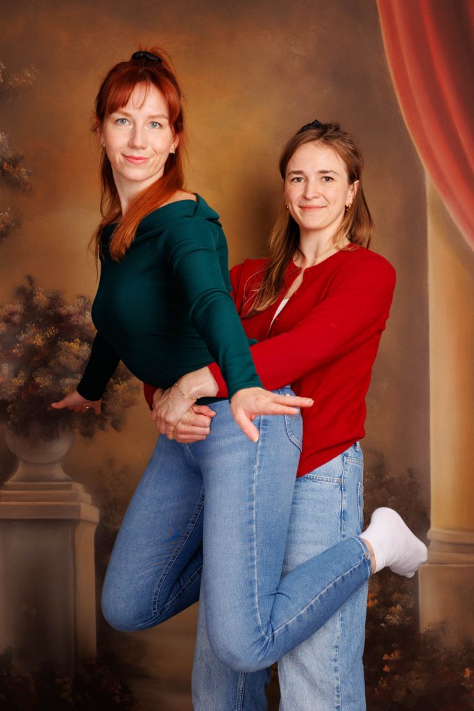 Two women posing joyfully against a decorative backdrop with curtain and columns.