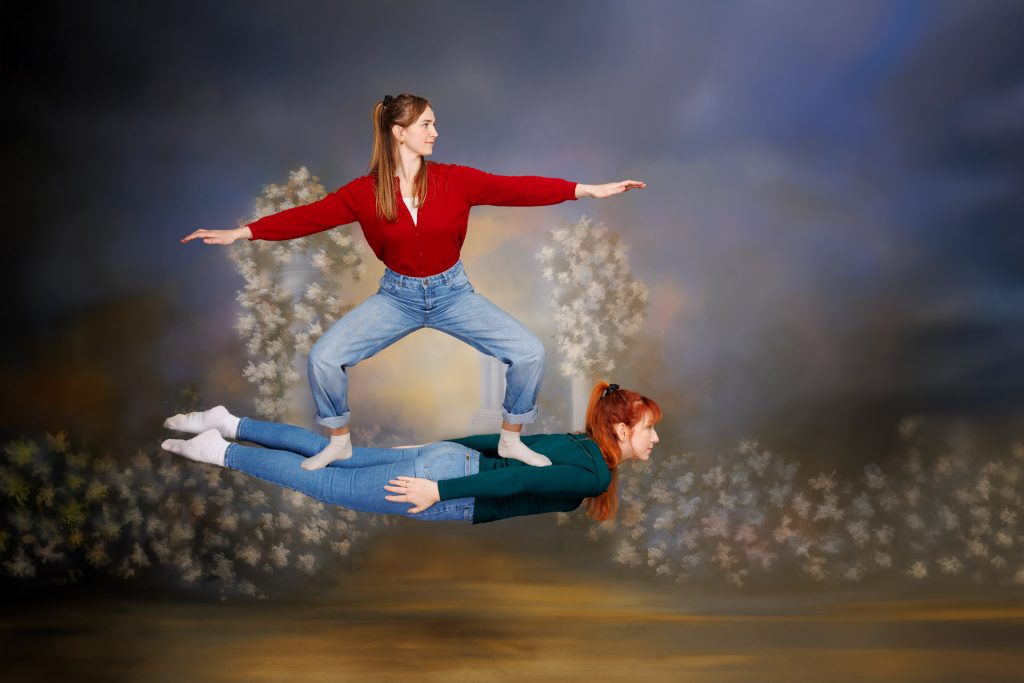 Two women in artistic pose against painted tree and column backdrop, one standing on the other.