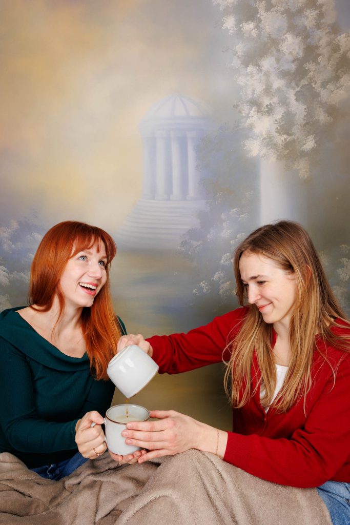 Two women with a cup and jug, blanket-covered, painted columns and trees in the background.