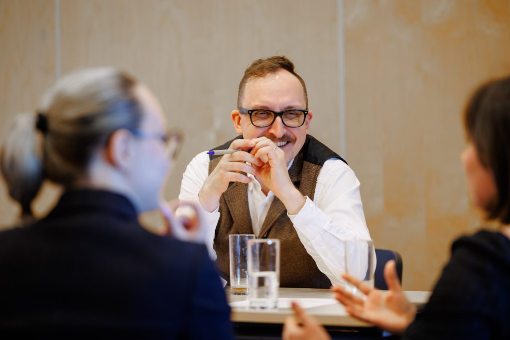 Uśmiechnięty mężczyzna w okularach i kamizelce słucha rozmówców przy stole na konferencji Stan Młodych 2025.
