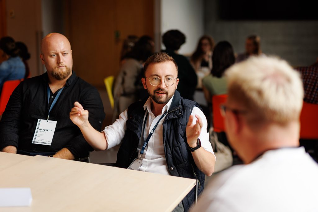 Mężczyzna z identyfikatorem gestykuluje podczas rozmowy przy stole na konferencji Stan Młodych 2025.