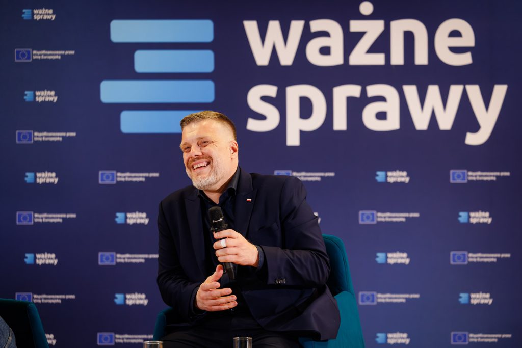 Adrian Zandberg z mikrofonem siedzi na scenie konferencji Stan Młodych 2025, na tle ścianki Ważne Sprawy.