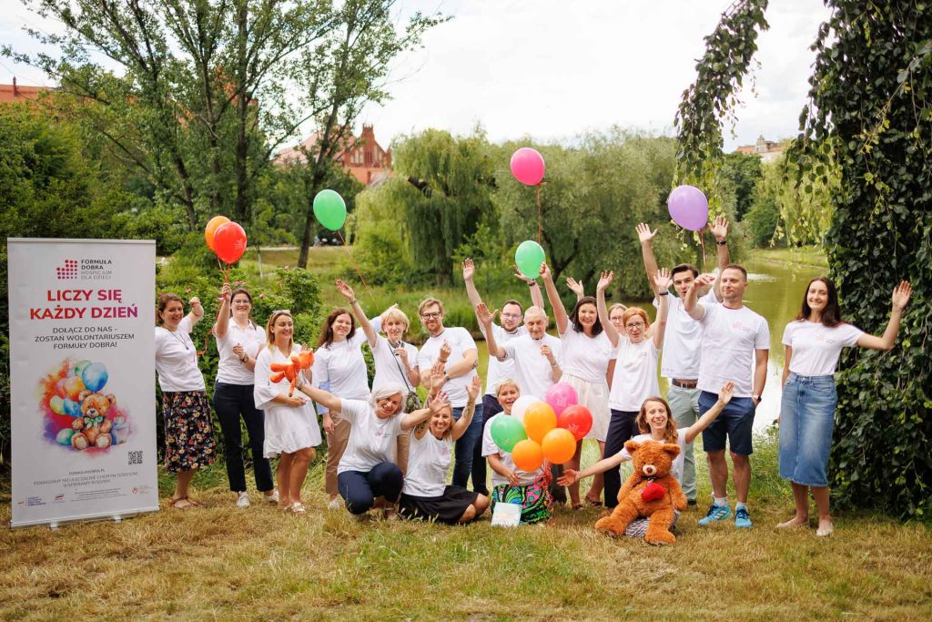Grupa osób z balonami i pluszakami stoi na trawie przy jeziorze obok roll-upa hospicjum dziecięcego.