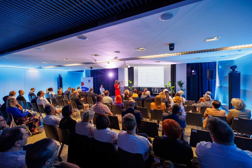 Sala konferencyjna w niebieskim świetle, publiczność słucha prelegentki.