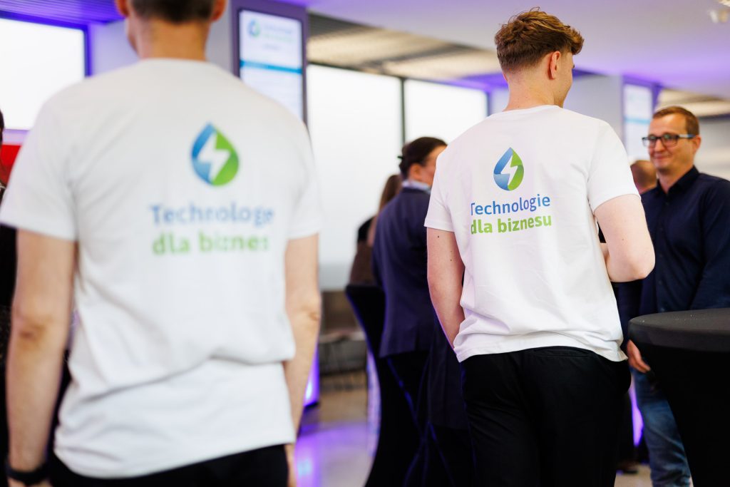 Osoby w koszulkach Technologie dla Biznesu rozmawiają z uczestnikami konferencji.
