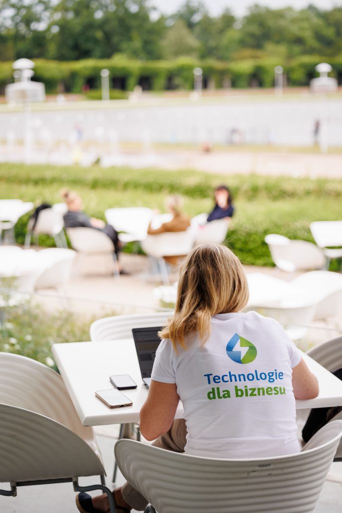Wolontariuszka w koszulce Technologie dla Biznesu siedzi na tarasie z widokiem na pergolę.