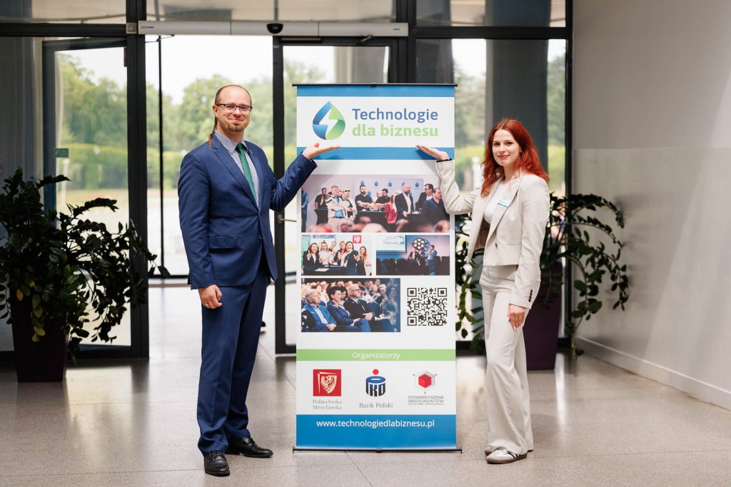 Kobieta i mężczyzna pozują przy roll-upie konferencji Technologie dla Biznesu.