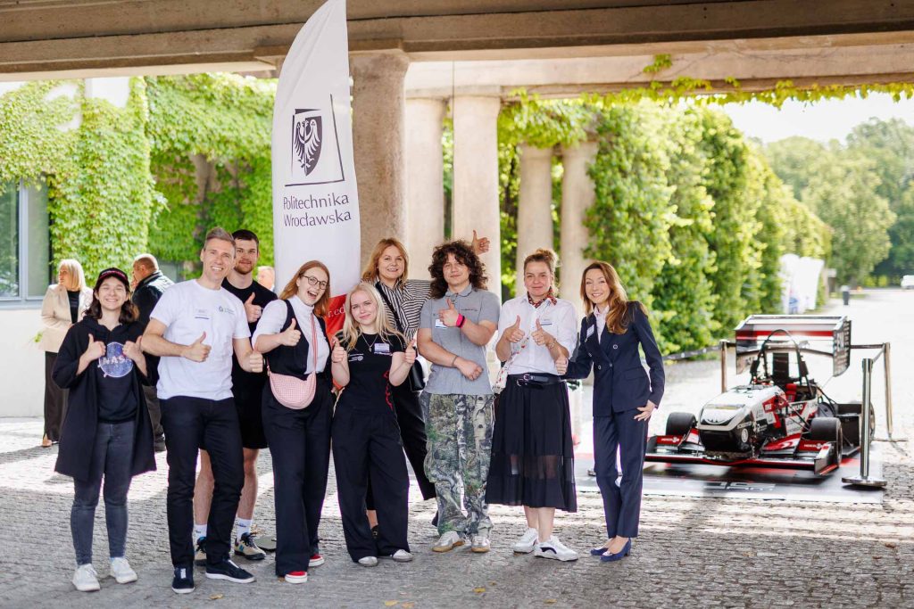 Grupa studentów pozuje obok bolidu wyścigowego i flagi Politechniki Wrocławskiej podczas konferencji Technologie dla Biznesu.