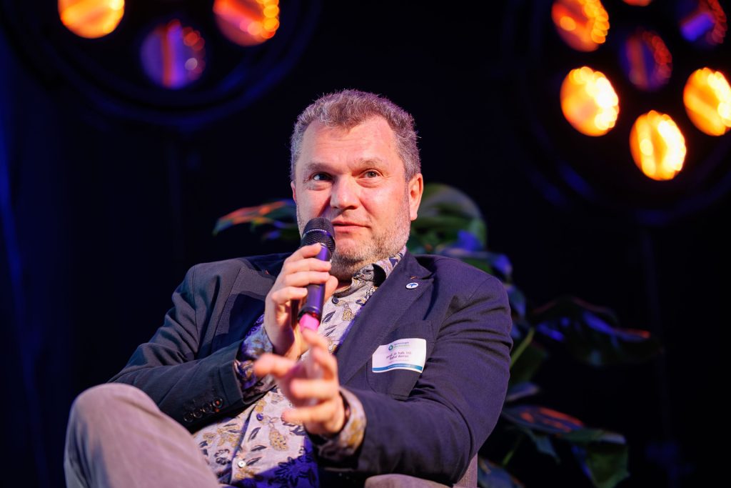 Panelista z mikrofonem mówi, siedząc w fotelu w kolorowym świetle sceny Technologie dla Biznesu.