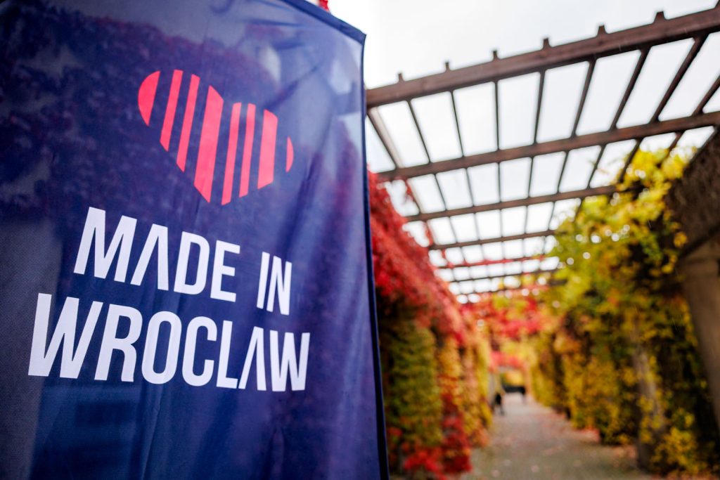 Granatowy baner Made in Wrocław obok pergoli porośniętej jesiennymi liśćmi.