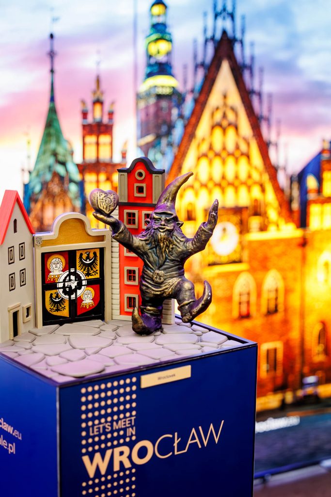 Statuetka z wrocławskim krasnalem na tle kolorowej ilustracji ratusza – nagroda Made in Wrocław 2025.