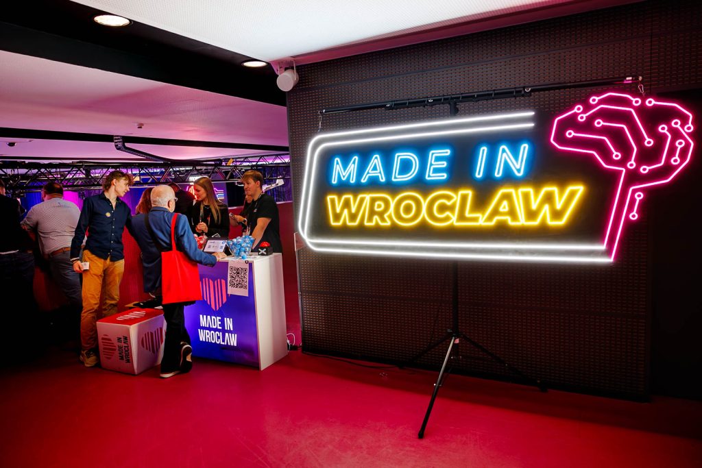 Neon Made in Wrocław przy stoisku, przy którym rozmawiają uczestnicy.